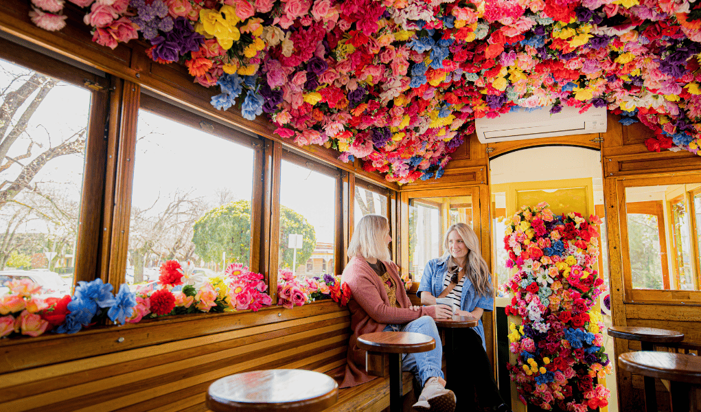 Two women sitting inside a tram, are talking and drinking coffee. the tram's ceiling is covered in flowers of pinks, purples, oranges, and yellows, the flowers also run along the frame of the tram's windows